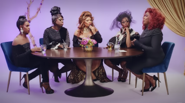Five People Sit At Round Table, All Are Black Drag Queens In Black Dresses And Outfits. Purple Hue Background.